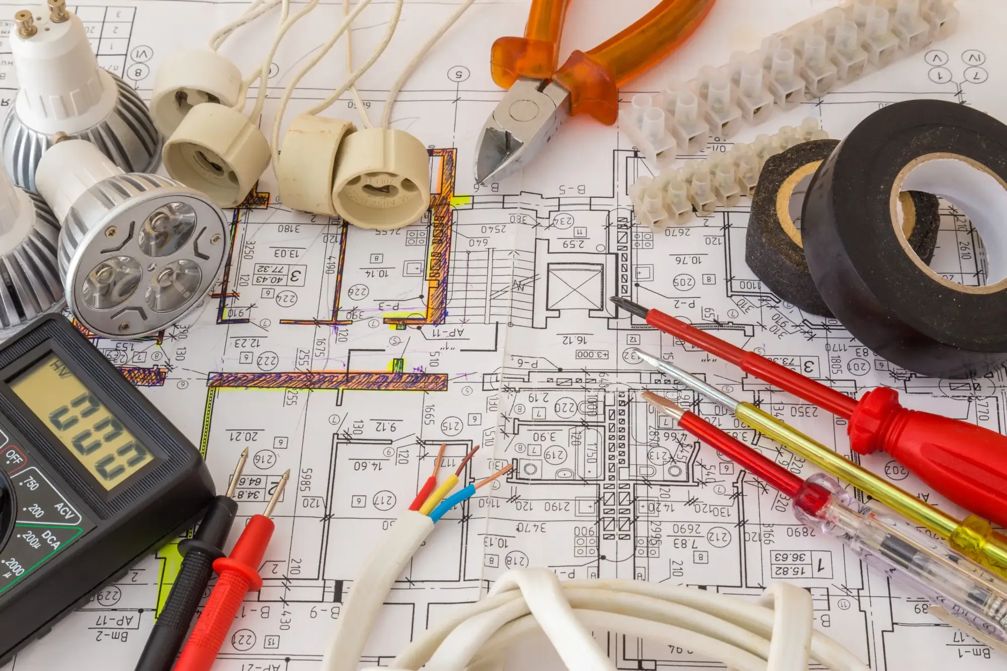 A collection of electrical tools, including LED light bulbs, a digital multimeter, wire cutters, electrical tape, and screwdrivers, placed on top of a detailed building floor plan with marked electrical wiring routes.