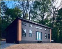 A tiny house painted dark with an angled roof on the edge of a forested area